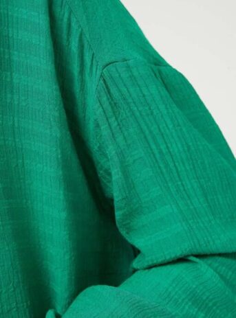 Camisa amplia en tejido crepe con textura de color verde. Con cuello solapa y manga larga terminada en puño. Modelo con el bajo trasero ligeramente más largo. De compañía Fantástica